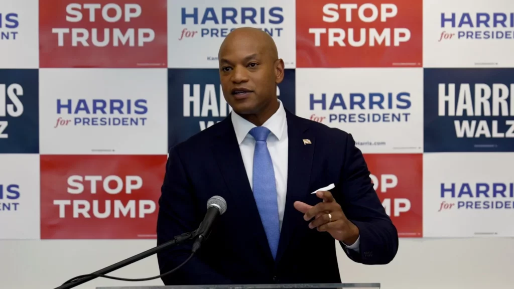 Gov. Wes Moore answering questions at press conference with "Harris/Walz" and "Stop Trump" signs behind him.