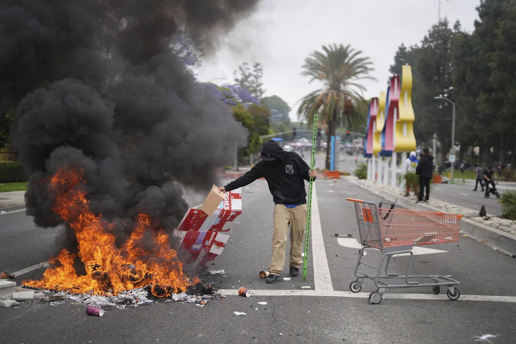 A protester places debris in a fire as Border Patrol personnel in riot gear and gas masks stand guard outside an industrial park in Paramount, Calif., on Saturday, June 7, 2025.