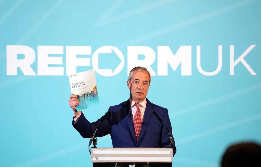 Reform UK leader Nigel Farage holds up a piece of paper as he speaks in front of a blue Reform UK sign.