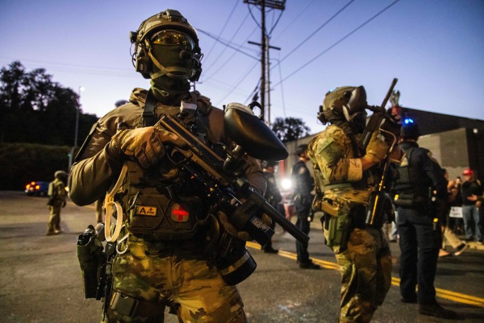 Federal officers equipped with weapons and gear stand at the ready near an ICE facility in Portland.